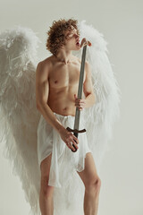 Young man with curly hair and wings poses with sword from which he eats raw meat against white studio background. Concept of fashion and beauty, modern art and historical fiction fusion. Ad