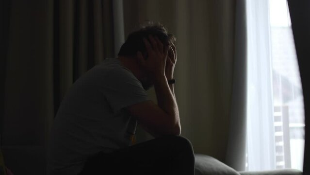 Distraught man sitting on a sofa, face buried in hands and rocking restlessly, with room darkness illuminated by window light.