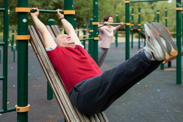 Senior man goes in for sports in city park on sports ground - doing exercises to pull his legs to chest