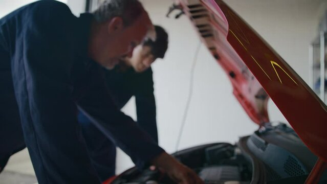 Experienced car mechanic servicing car engine looking under hood with male trainee - shot in slow motion