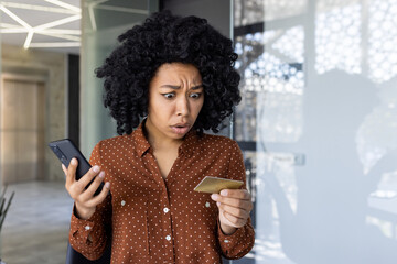 A young woman looks shocked while examining her credit card and smartphone. The setting suggests a...