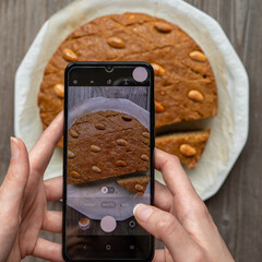 Woman hands take a smartphone photo of a helbeh cake (fenugreek cake). Phone photography of sweets for a blog or social media. Homemade round cake with almonds, fenugreek seeds, and semolina.