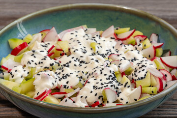 Red radish and cucumber salad with yogurt dressing and nigella seeds in a bowl standing on rustic wooden boards. 