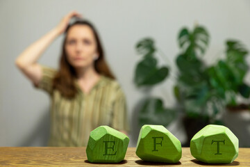 Letters EFT written on green painted wooden blocks. Female tapping  top of head (TH) meridian point in blurred background. Emotion-focused therapy treatment concept.