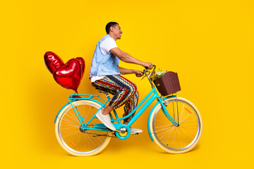 Full size photo of cheerful nice man dressed denim vest ride cycle with balloons look empty space isolated on yellow color background