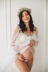 Smiling pregnant woman  in white polka dot peignoir and gypsophila wreath posing near white wall in...