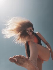 A young woman with long blond hair smiles and laughs while kicking her foot in the air. She is...