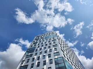 Impressive modern architecture of a tall office building with sleek glass facade and unique geometric design reaching high into the sky against a backdrop of vibrant blue sky and fluffy white clouds
