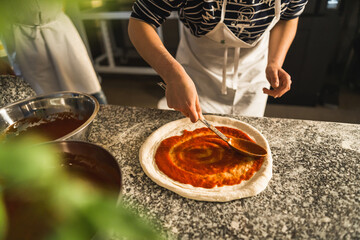 Hands adding fresh tomato sauce to pizza dough. Pizza ingredients on the wooden table. High quality...