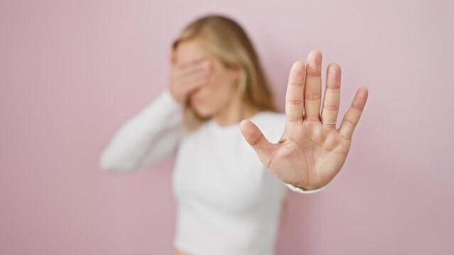 Terrified young blonde woman, covering eyes with hand, making stop gesture in fear over pink isolated background. embarrassing, negative expression signifying problem.
