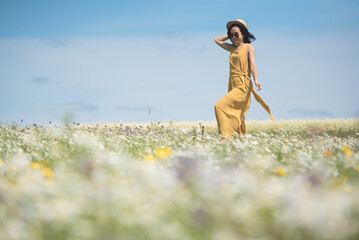 A young Asian woman wearing a yellow jumpsuit dress and sunglasses holds her straw hat as she walks across a wild flower meadow in a sunny summer day in Kinross, Scotland, UK