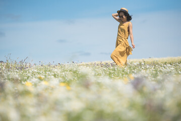 A young Asian woman wearing a yellow jumpsuit dress and sunglasses holds her straw hat as she walks...