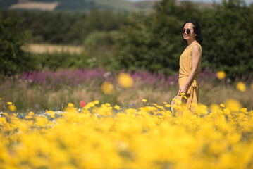 A young Asian woman wearing a yellow jumpsuit dress and sunglasses walking elegantly across a wild yellow flower meadow in a sunny summer day in Kinross, Scotland, United Kingdom