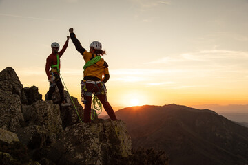 Two people are standing on a mountain top, one of them is holding a hand up in the air