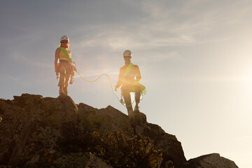 Two people are standing on a rocky mountain, one of them holding a rope. The scene is bright and...