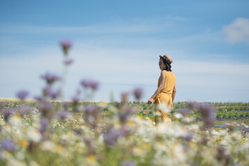 A young Asian woman wearing a yellow jumpsuit dress and a straw hat walks elegantly across a wild...