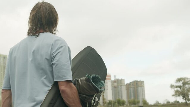 Medium back view shot of long-haired male wakeboarder carrying board with boots attached to bindings while going towards water on cloudy day