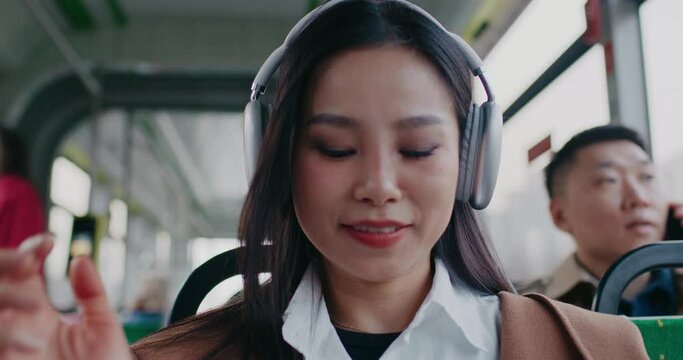Facial portrait of Asian girl commuting in public transport. Passengers in blurred background. Woman putting on large new wireless headphones to enjoy good quality music. Using technology gadgets.