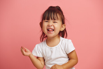 Cheerful mischievous little asian appearance girl shows her tongue to the camera, smiling cheerfully while standing on a pink polished background. Joyful child headshot portrait.