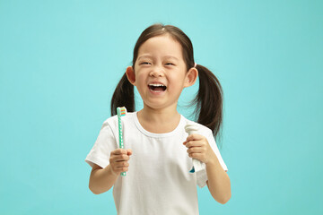 Cheerful little asian ethnicity girl holding squeezed toothpaste tube and toothbrush, fun laughing, wearing in white t shirt, standing over blue isolated background. - 795251456