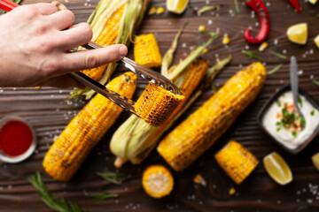 Kitchen tongs with grilled corn on the cob pieces in caucasian hands on kitchen wooden table...