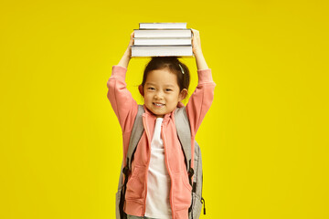 Sweet first grader asian ethnicity schoolgirl holding books above head, cheerful smiling standing with backpack on her shoulders over yellow isolated background. - 795252075