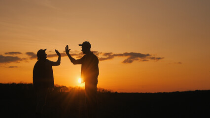 Silhouettes of two farmers in a high five gesture. Standing in a field at sunset