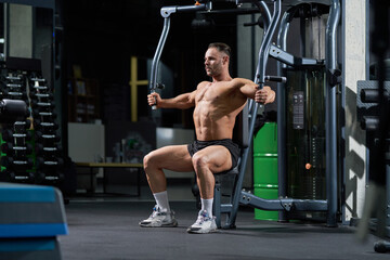 Shirtless male athlete wearing black shorts, training in gym. Side view of fit sportsman doing pull-downs, lifting metal plates of cable crossover machine, copy space. Sport, weightlifting concept.