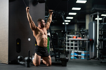 Serious attractive sportsman doing exercise for biceps, standing on knees in gym. Side view of muscular male athlete in black shorts, pulling cables of training apparatus, looking away. Sport concept.