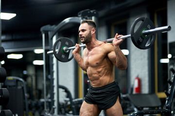 Determined, strong athlete in black shorts, holding heavy barbell in gym. Side view of muscular bearded bodybuilder keeping barbell behind back, while training. Bodybuilding, weightlifting concept.