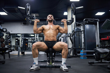 Fototapeta na wymiar Attractive bearded man doing exercise with training apparatus in gym. Low angle view of strong male athlete sitting, using sport equipment for building muscles indoors. Sport, weight lifting concept.