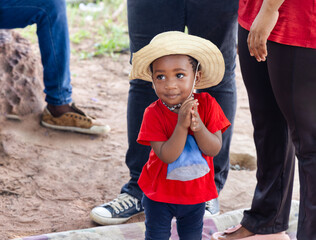 small african child girl in front of the house in the yard, wearing a red t-shirt and a straw hat