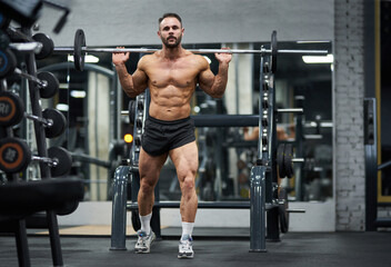 Determined, strong athlete in black shorts, holding heavy barbell in gym. Full body of bearded bodybuilder keeping barbell behind back, while exercising indoors. Bodybuilding, weightlifting concept.