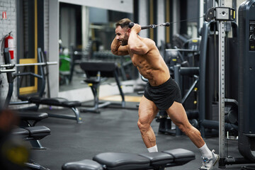 Strong Caucasian athlete training in crossover machine in gym. Side view of healthy bearded sportsman doing pull-downs, lifting metal plates of cable crossover machine, copy space. Crossfit concept.