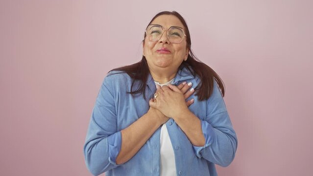 Cheerful middle age hispanic woman, standing with hands over heart, eyes closed in gratitude, radiates health. beautifully isolated over pink background, affirming a joyful, confident lifestyle.
