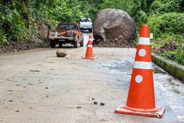 selective focus Orange cones on the road Large boulders fall from the mountain, blocking a rural concrete road in a green forest during the rainy season. Cars drive through the narrow lane.