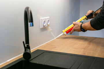 Glazier is using a silicone caulking gun to fill gap between glass backsplash and countertop in new modern kitchen - obrazy, fototapety, plakaty