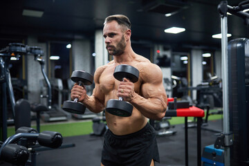 Determined handsome male weightlifter working out in gym. Portrait of bearded man with hairstyle, lifting dumbbells and looking aside, against blurred background. Bodybuilding, sport concept.
