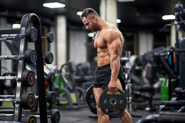 Strong man in sportswear lifting plates for barbell in gym. Side view of muscular sporty man carrying round plates, going to exercise, against blurred gym background. Concept of weightlifting, sport. 
