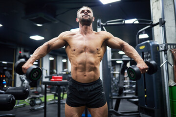 Shirtless Caucasian bodybuilder working out chest muscles, while using dumbbells. Low angle view of male crossfit athlete exercising in modern gym at evening. Concept of sport, bodybuilding.