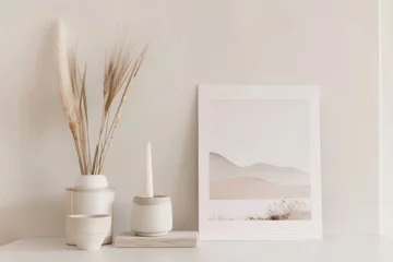 Deurstickers A minimalist still life image of a ceramic vase with wheat stalks, a ceramic candle holder with a candle, and a framed print of a desert landscape. The objects are arranged on a white table against a © Pachara