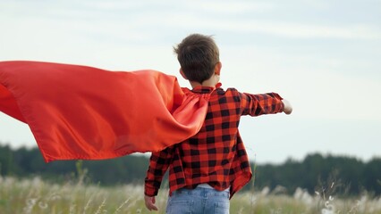 Boy plays superhero in red cape, childhood dream. Happy child playing superhero against sky. Little hero in red cloak watches sunset. Brave child winner in red raincoat plays in nature. Kid dreams