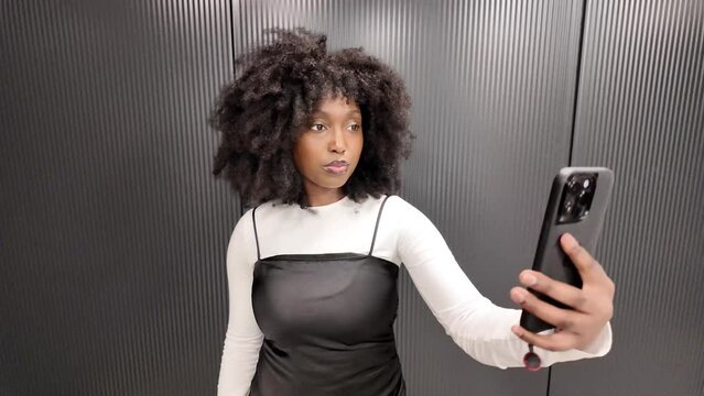 A young African American woman stands inside an elevator, engaging with her smartphone. In the first instance, she is focused on reading, possibly browsing or texting, capturing the essence of modern