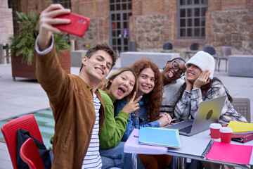 Group cheerful international students taking funny selfie photo sitting at table in university campus. Diverse joyful young people looking at front camera of mobile phone and smiling together outdoor