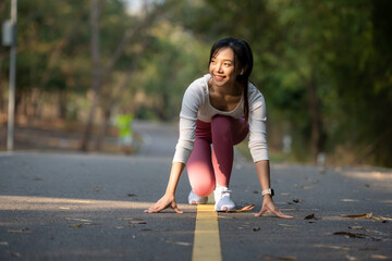 A focused female athlete in sportswear crouches at the starting line on a park path, ready to...