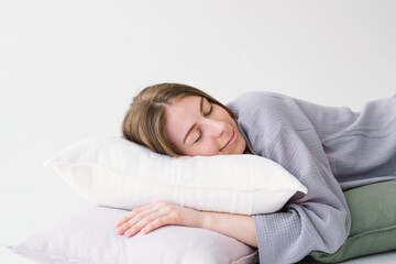 A woman lies comfortably on her side, embracing a white pillow with a tranquil expression. She is wearing a pajamas and resting against a light, solid-colored backdrop, evoking a sense of relaxation