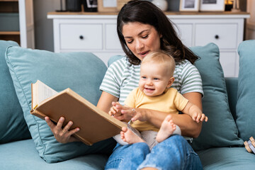 Young happy mother reading a fairytale story book to her baby. Mommy and kid sitting on sofa at...