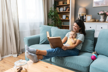 Young happy mother reading a fairytale story book to her baby. Mommy and kid sitting on sofa at home enjoying in imagination. Parent and child lifestyle.
