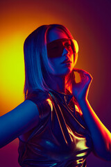 Obraz na płótnie Canvas Futuristic urban chic. Portrait of beautiful young girl n sunglasses and metallic dress against gradient orange yellow background in neon light. Concept of modern fashion, trends, beauty, youth