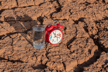 A glass of drinking water and an alarm clock against the backdrop of heat-cracked clay in the desert